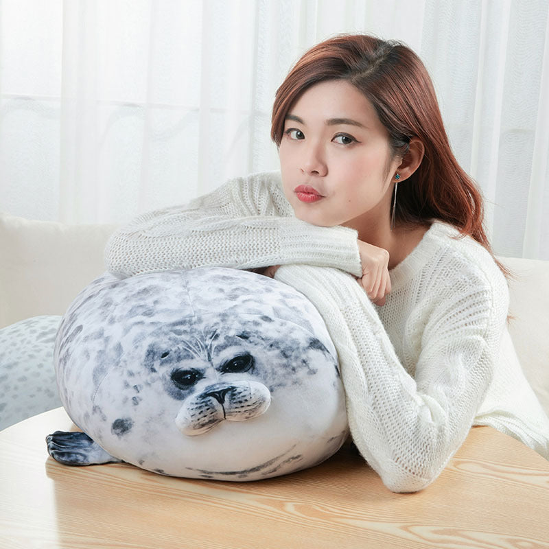A woman is resting her chin on a large, round, gray and white seal plush pillow.