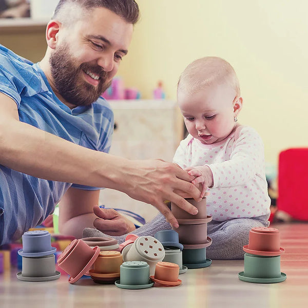A man is helping a toddler stack colorful silicone cups on a wooden table.