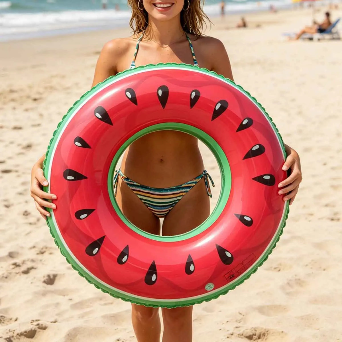A woman holds a bright red inflatable fruit ring float on a sandy beach.