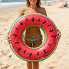A woman holds a bright red inflatable fruit ring with black seeds on a sandy beach.