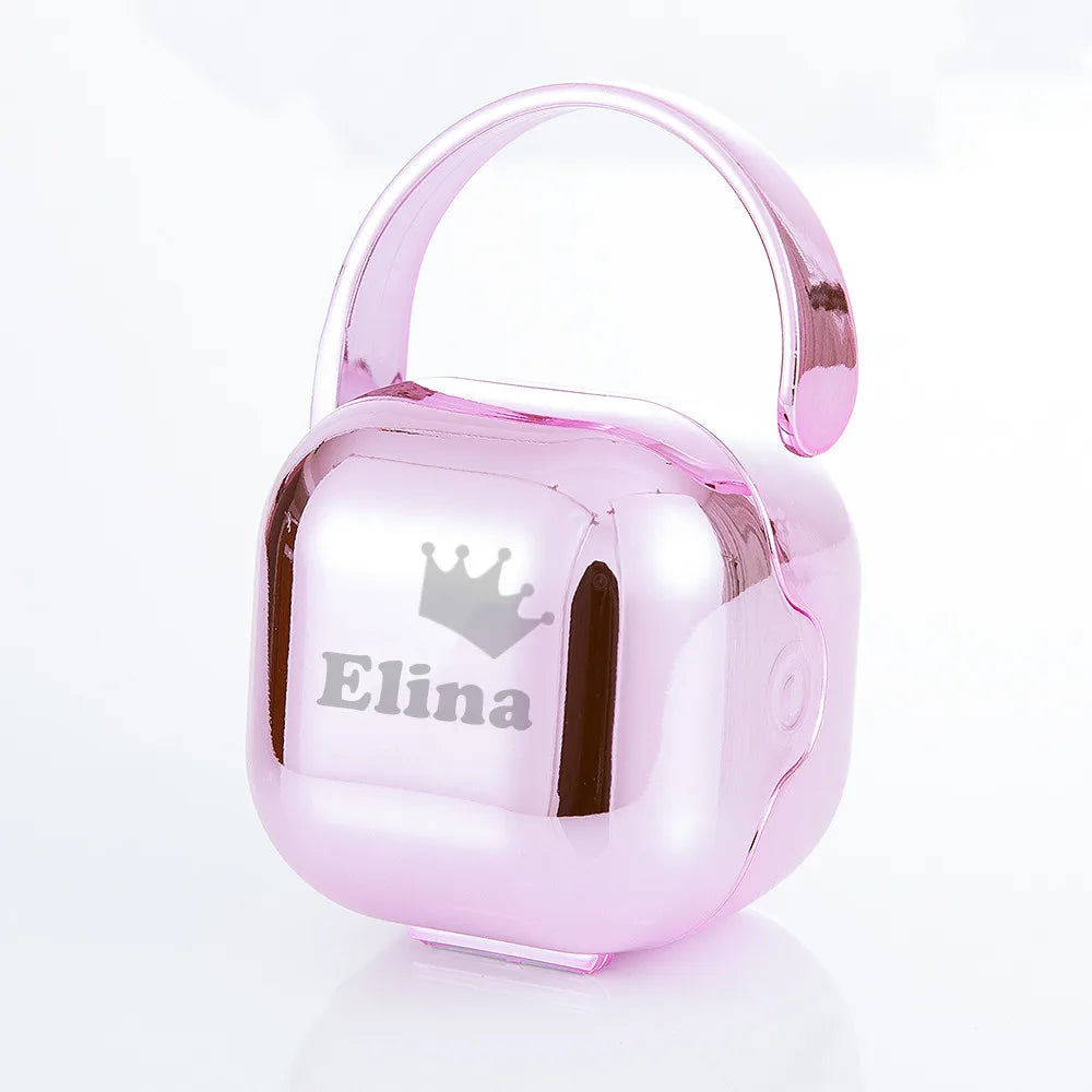 A pink silicone baby pacifier with a crown design stored in a clear plastic case.