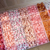 Assortment of pink, red, and white acrylic beads for jewelry making with gold accents displayed in clear plastic trays.