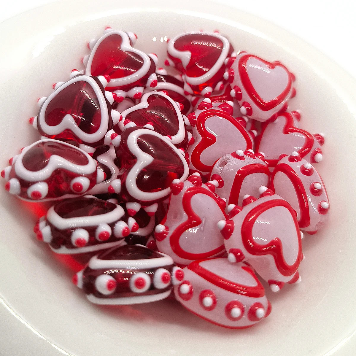 A pile of red and pink heart-shaped lampwork glass beads for jewelry making arranged in a white bowl.