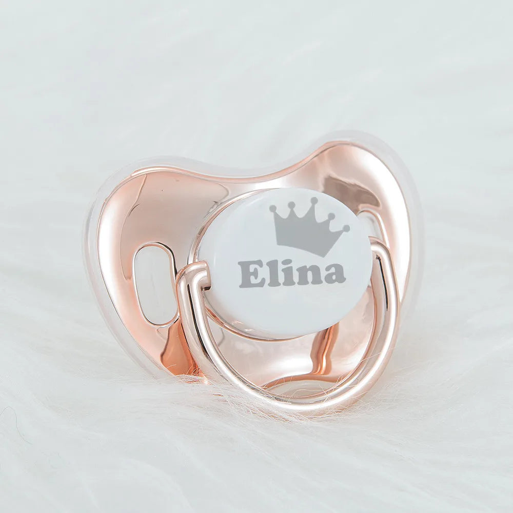 A personalized baby pacifier with a crown design and storage case in rose gold metal.