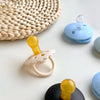 A natural latex baby pacifier in yellow and white is shown next to a woven mat and blue silicone components.