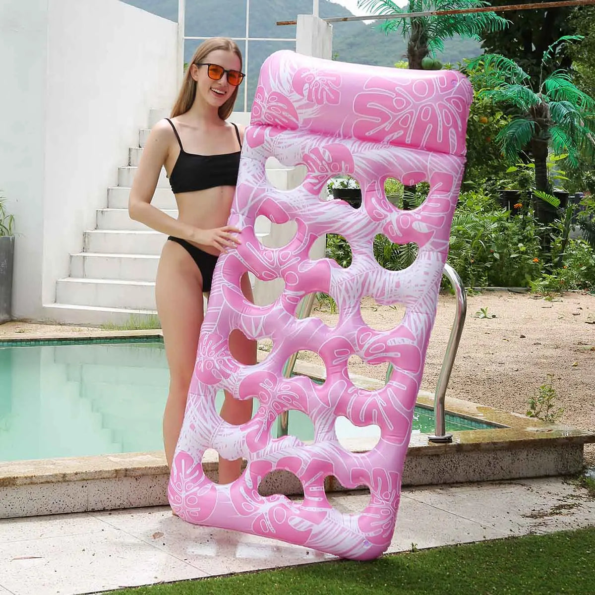 A woman stands next to a pink inflatable pool float with a headrest in a backyard setting.
