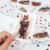 A hand holds a playing card featuring a vintage illustration of a cat with a sword, surrounded by other cards.