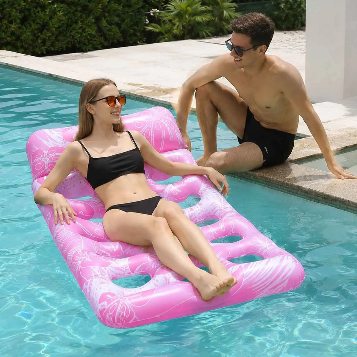 A woman relaxing on a pink inflatable pool float in clear blue water with a man nearby.