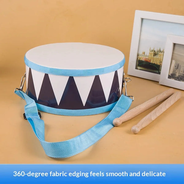 A white fabric drum toy with a blue edge and a strap is shown next to wooden sticks and a framed picture.