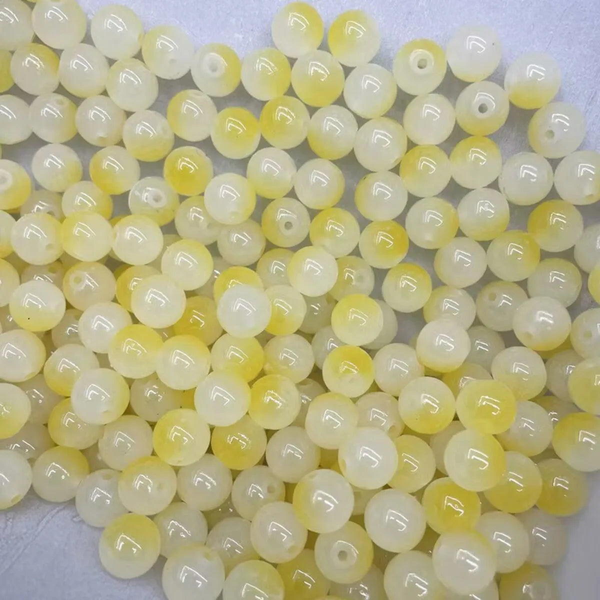 A close-up view of a 50-pack of dual-color glass round beads, featuring pale yellow and translucent white shades.