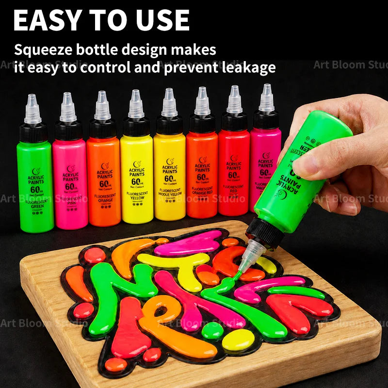 A hand squeezes a neon green bottle of acrylic paint onto a wooden board with colorful paint designs.