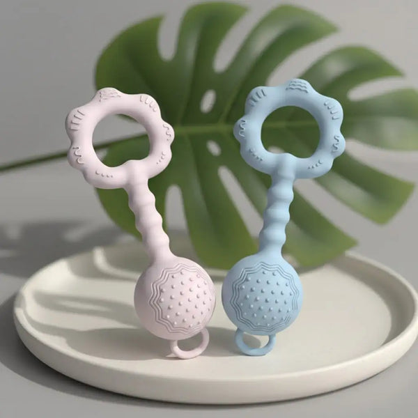 A silicone baby rattle and teether set in pink and blue resting on a light gray plate with a green leaf background.