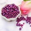 A bowl filled with assorted purple and magenta pearl beads next to a pink rose petal.