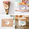 A hand is placing a round thank you sticker on a gift box, surrounded by pink roses and natural wood.
