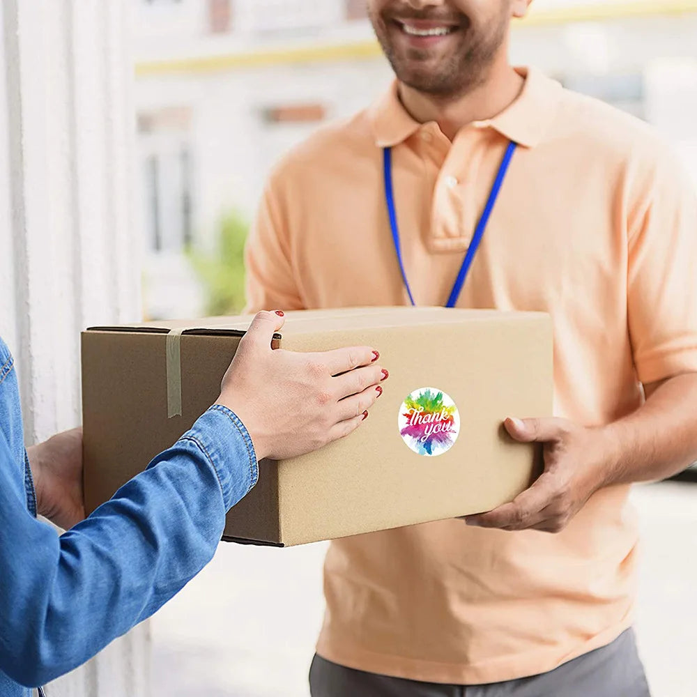 A man in an orange shirt is handing a cardboard box to another person, with a colorful thank you sticker visible on the box.