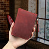 A hand holds a deep red leather coin purse with a black zipper, set against a rustic brick and window background.