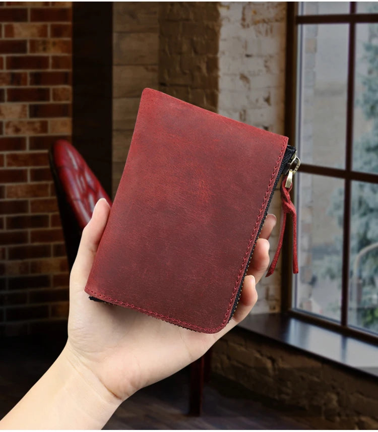 A hand holds a deep red leather coin purse with a black zipper, set against a rustic brick and window background.