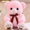 A pink plush teddy bear with black eyes and a brown bow sitting on a wooden surface.