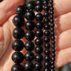 A hand holds multiple strands of dark, polished gemstone beads for jewelry making.