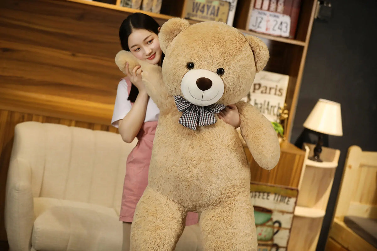 A young woman is hugging a large, light brown teddy bear plush toy in a cozy indoor setting.