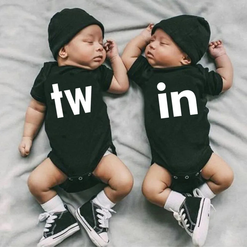 Two infants wearing black letter print bodysuits, one with 'tw' and the other with 'in', lying on a light gray surface.