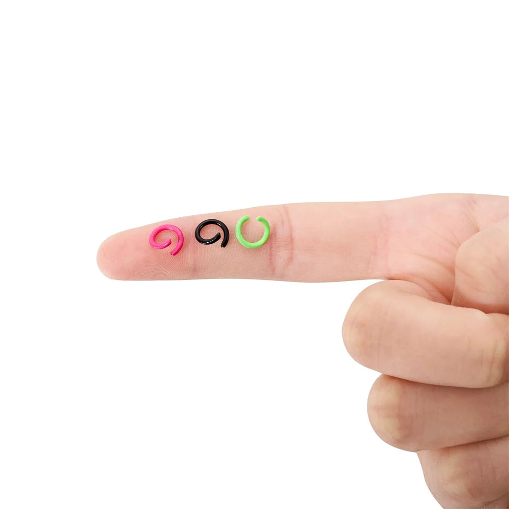 A hand holds three colorful metal open jump rings: pink, black, and green.