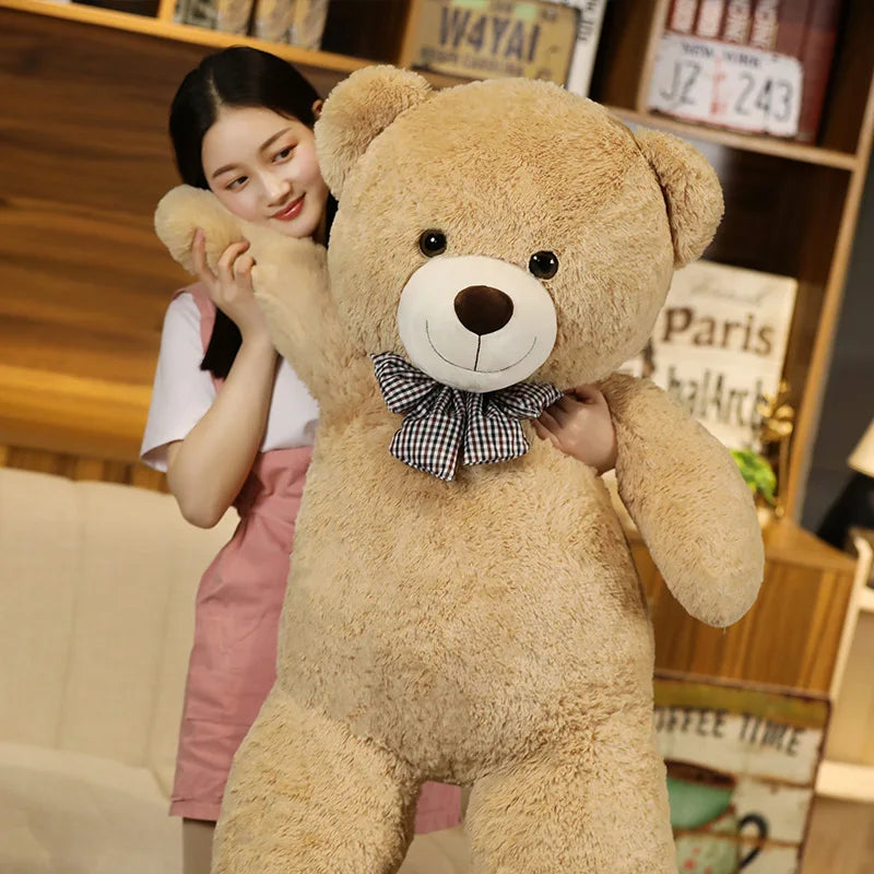 A young woman is holding a large, light brown teddy bear plush toy with a bow on its chest.