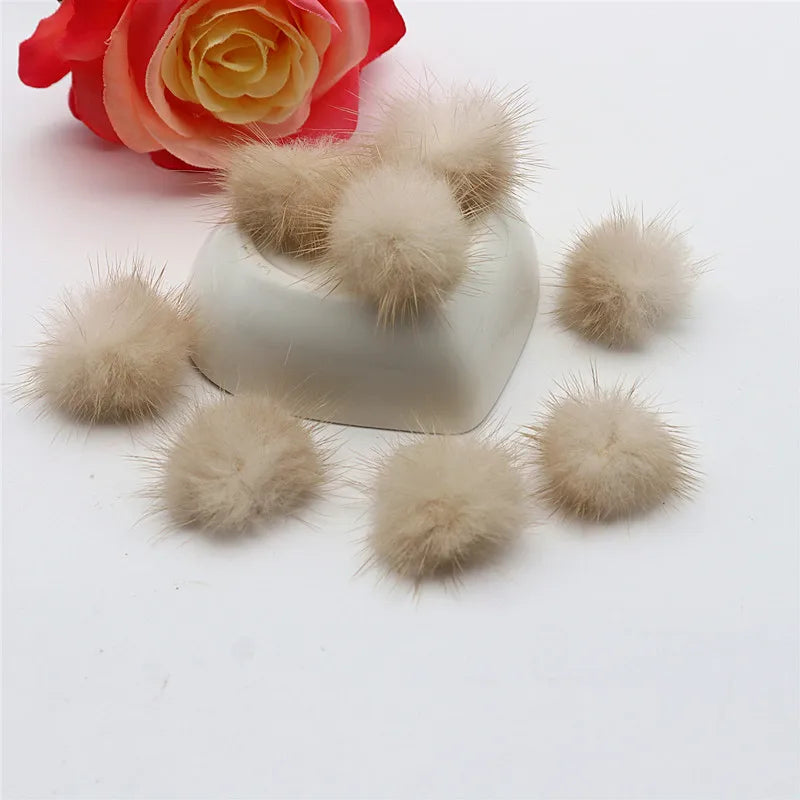 Several fluffy mink fur pom poms in various shades of beige and light brown are displayed next to a red rose.