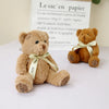 A soft brown teddy bear plush toy with a light yellow ribbon and a paper bag in the background.
