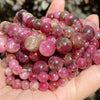 A hand holds a cluster of natural gemstone beads in shades of pink, purple, and reddish-brown.