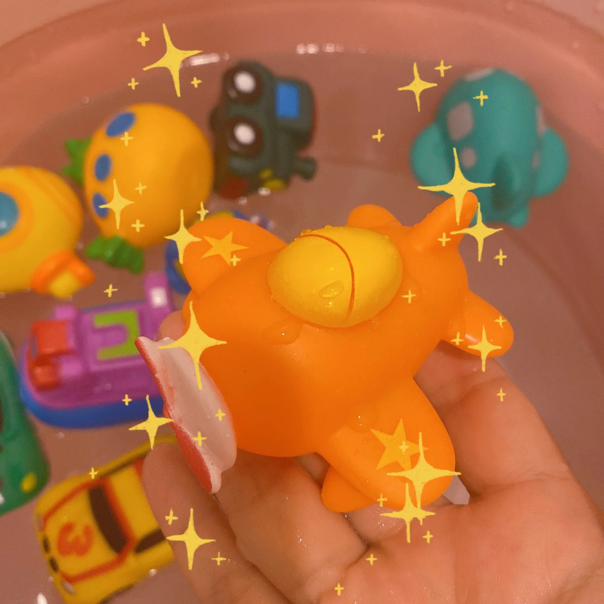 A hand holds an orange cartoon soft rubber bath toy resembling a stylized animal or vehicle in a pink bowl filled with water.