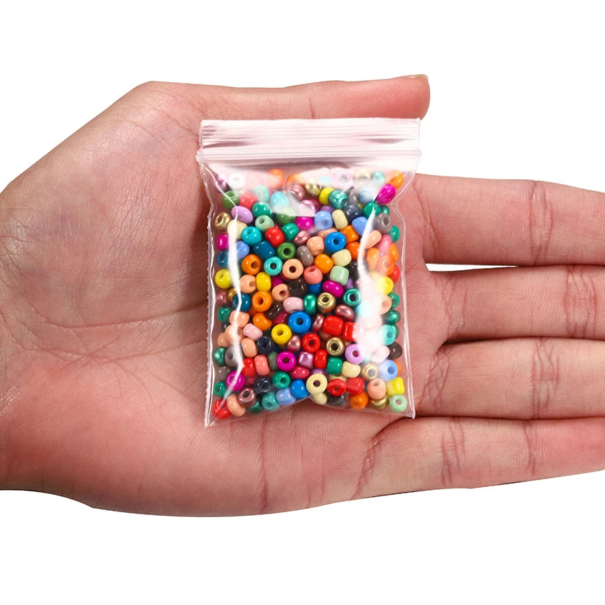 A hand holds a small clear plastic bag filled with various colored round glass seed beads for jewelry making