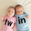 A baby wearing a pink letter print bodysuit is lying on a white surface.