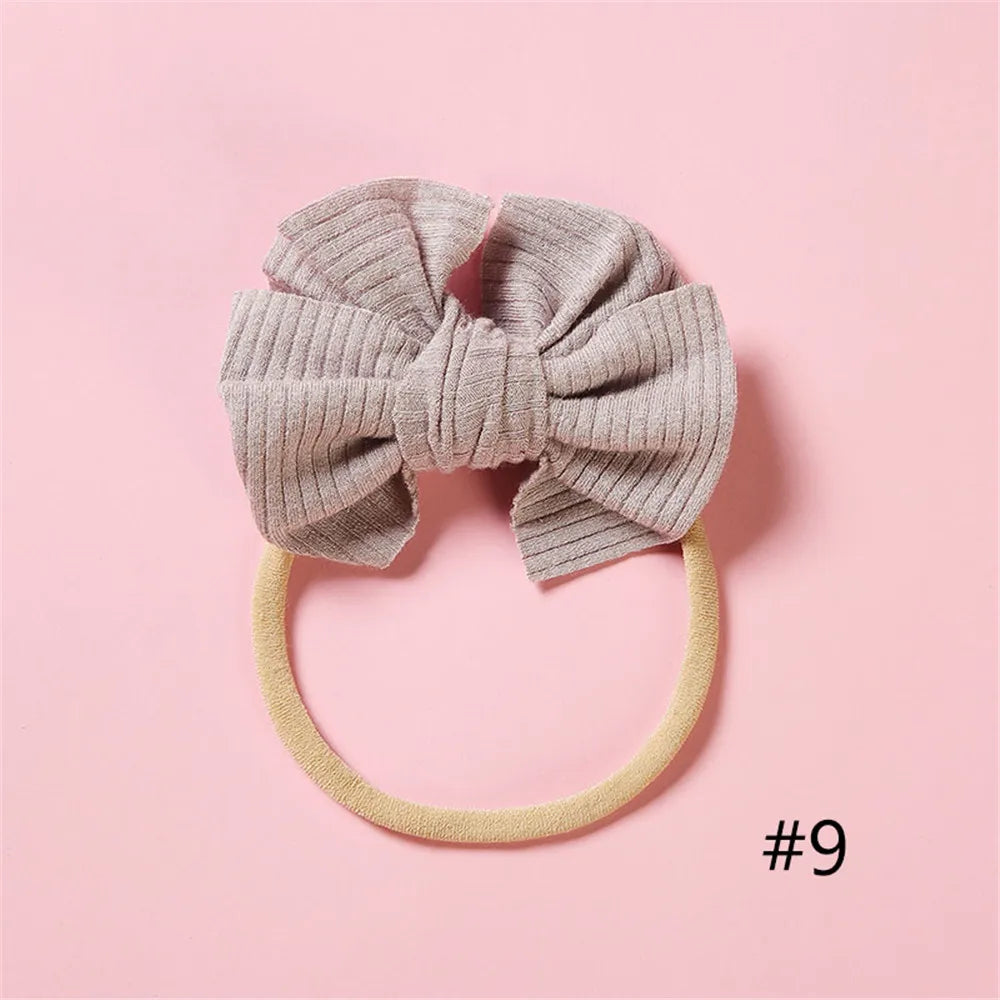 A beige ribbed fabric bow headband is displayed against a pink background.