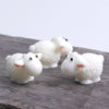 Three white miniature sheep figurines with pink ears resting on a dark wooden surface.