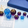 A collection of iridescent blue and purple resin glass spheres with gold glitter set against a light blue background.