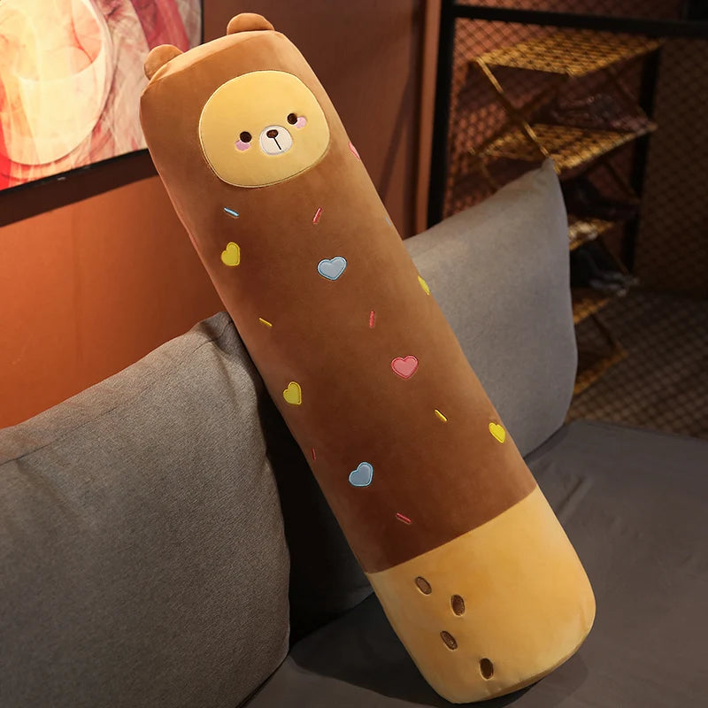 A long, cylindrical stuffed animal plush pillow resembling a bear, colored brown and yellow, with small colorful hearts scatt