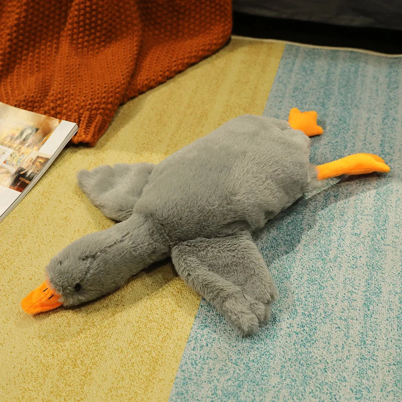 A gray oversized kawaii goose plush pillow with orange beak and feet resting on a light yellow and blue textured surface.