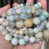 A collection of round natural gemstone beads in shades of blue, green, and beige are held in a person's hand.