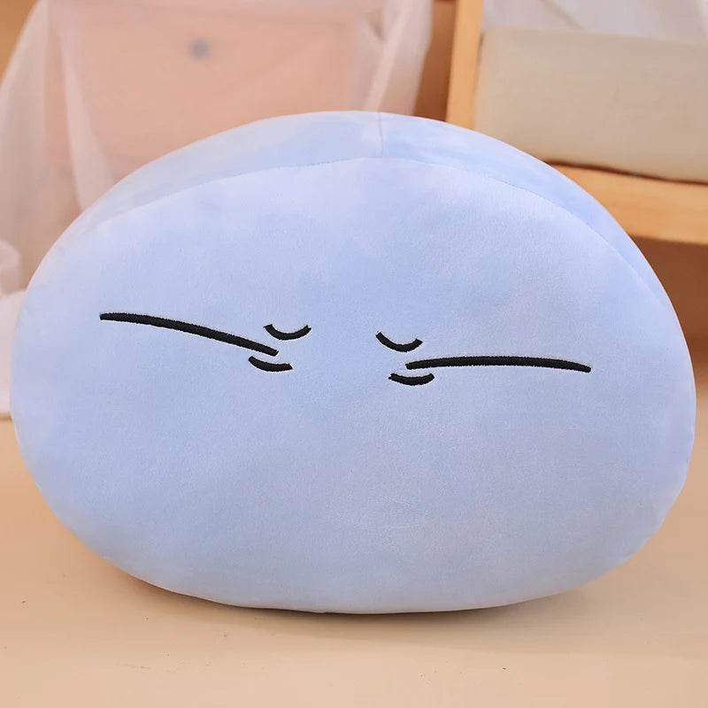 A light blue, round plush toy pillow resembling an anime slime character with closed eyes and simple lines for features.