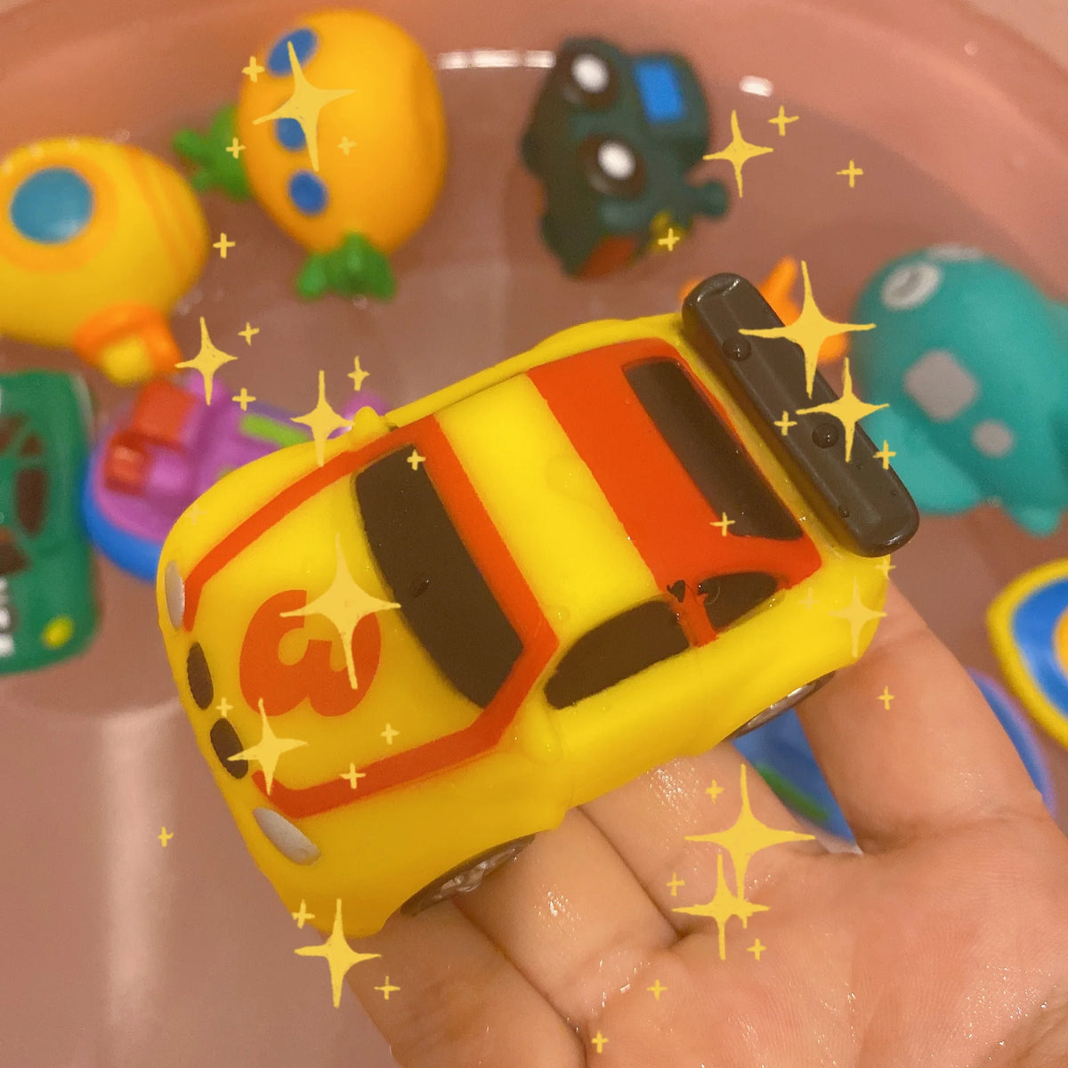 A hand holds a yellow cartoon soft rubber bath toy shaped like a vehicle, surrounded by other colorful toys.