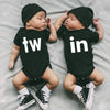 Two infants wearing black letter print bodysuits, one with 'tw' and the other with 'in', lying on a light gray surface.