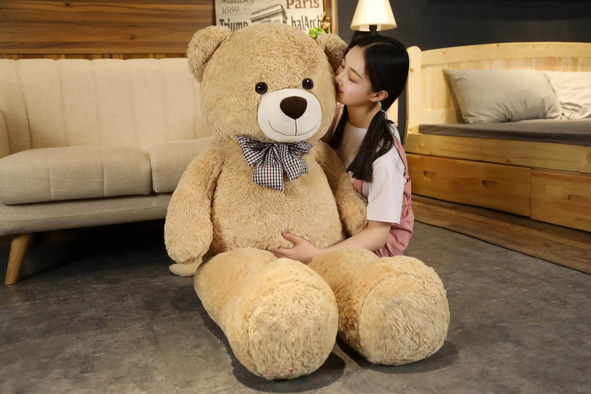 A young woman is sitting on the floor with a large, light brown teddy bear plush toy.