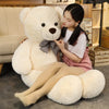 A young woman is sitting on the floor with a large white teddy bear plush toy and soft beige slippers in a wooden room.