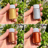 Three hands display metallic gold acrylic paint in glass bottles against a blurred green outdoor background.