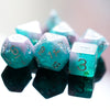 A close-up of a set of teal and green glitter resin polyhedral dice scattered on a reflective surface.