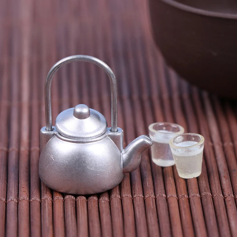 A small, silver miniature alloy kettle for dollhouse kitchen decor resting on a bamboo surface with a glass of liquid beside 