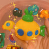 A hand holds a bright yellow rubber bath toy shaped like an animal, surrounded by other colorful toys.