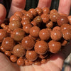A handful of small, round, reddish-brown gemstone beads with a sparkling finish is held in a hand.