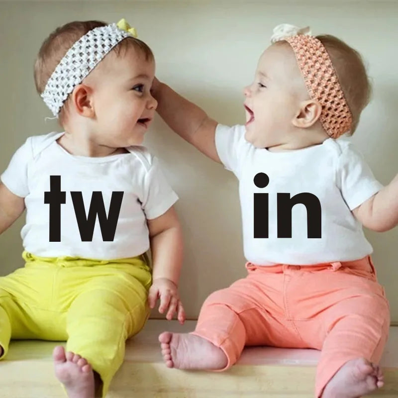 Two babies wearing white letter print bodysuits, 'tw' and 'in', sitting together.